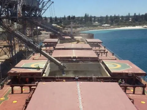 A bulk carrier with open hatches under a ship loader, next to the sea